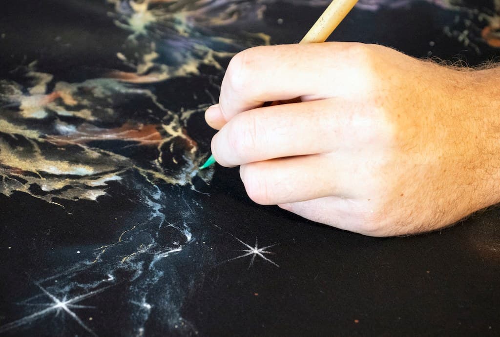 A brush painting a white star into the nebula on a black Galaxy shirt.