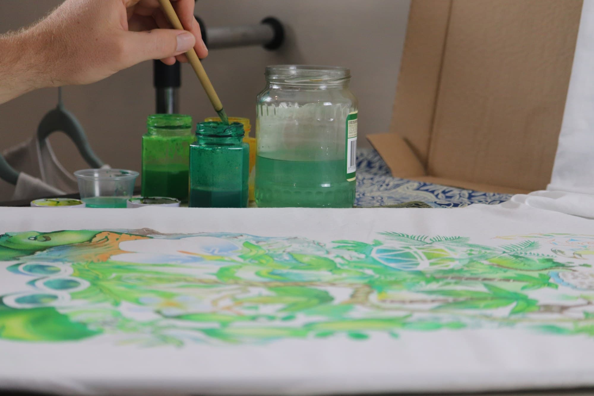 A brush and pots of green paint beside the Jungle shirt in progress.
