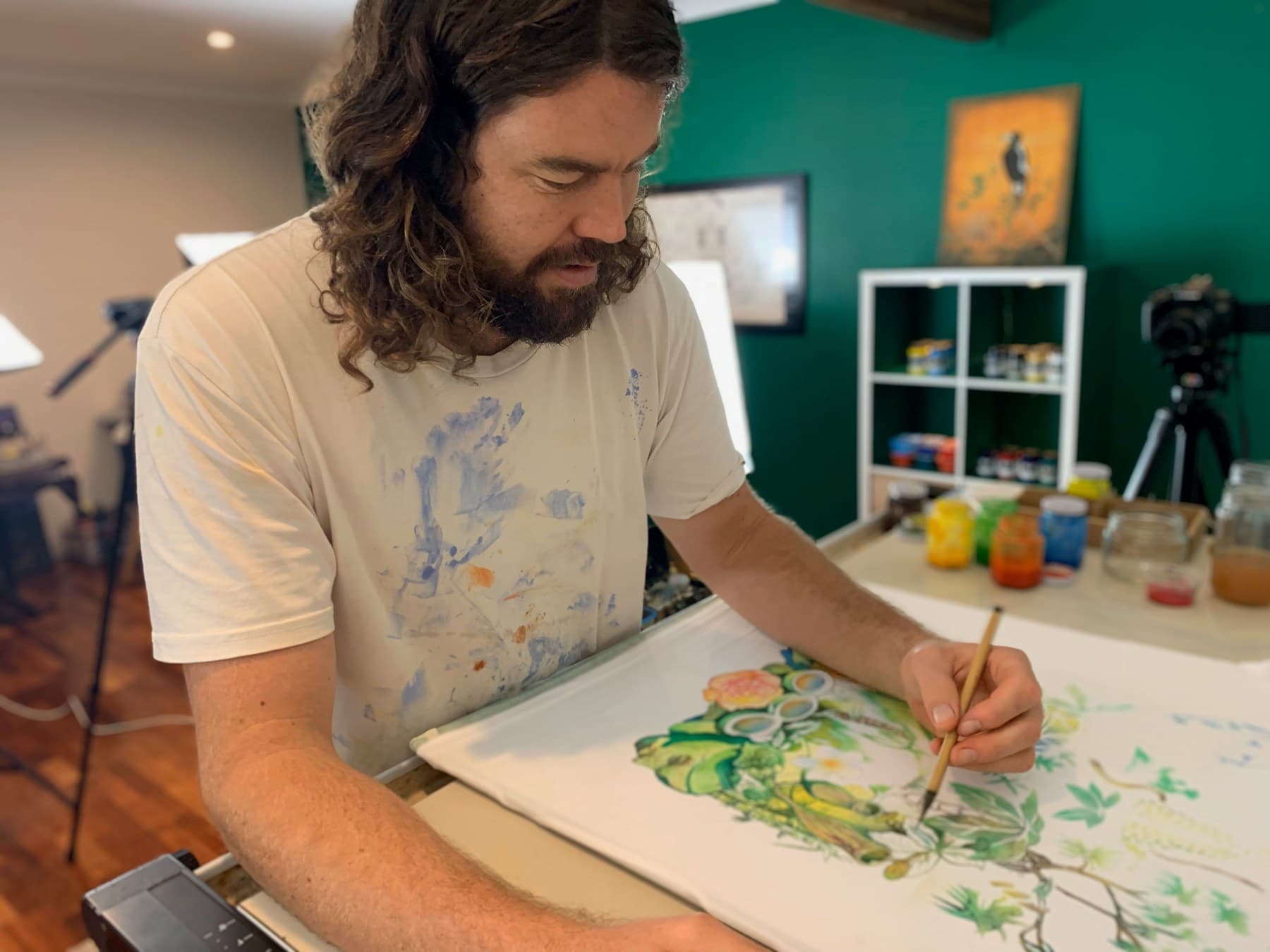 Sebastian painting a Jungle shirt at a table, with another shirt hanging behind him.