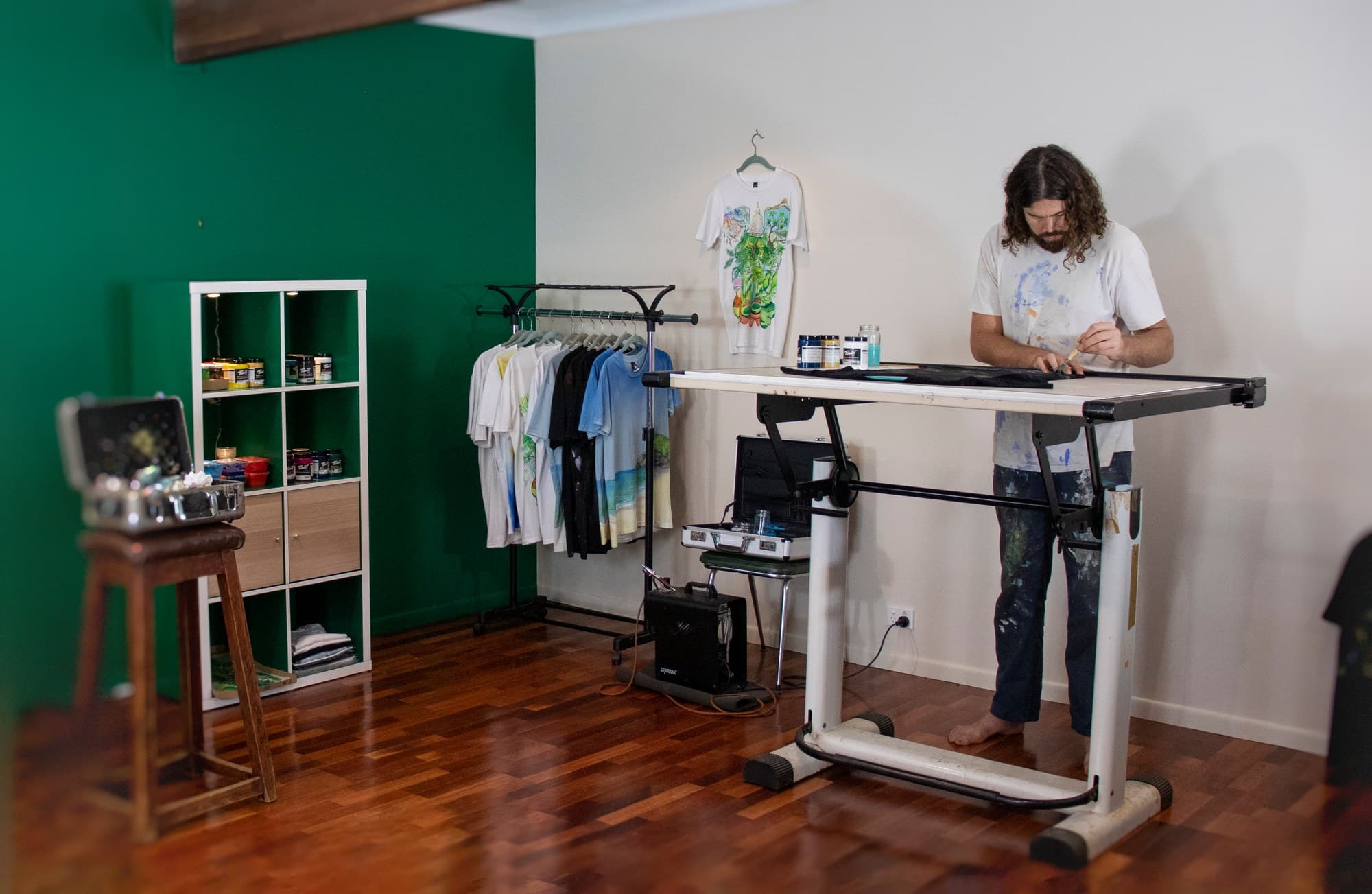 Wide studio view of Sebastian at the painting table, with shirts hanging beside him.